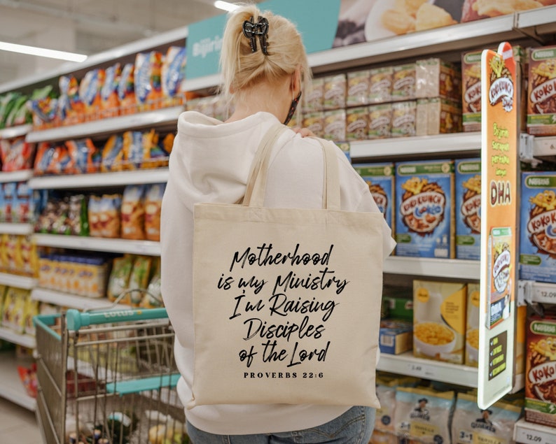 May include: A beige canvas tote bag with black text that reads "Motherhood is my Ministry I'm Raising Disciples of the Lord Proverbs 22:6". The bag is being carried by a person in a grocery store.