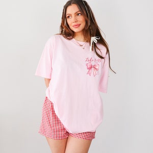 May include: An oversized pink t-shirt with a pink bow and the text "Luke 1:45" printed on the front. The model is wearing red and white gingham shorts.