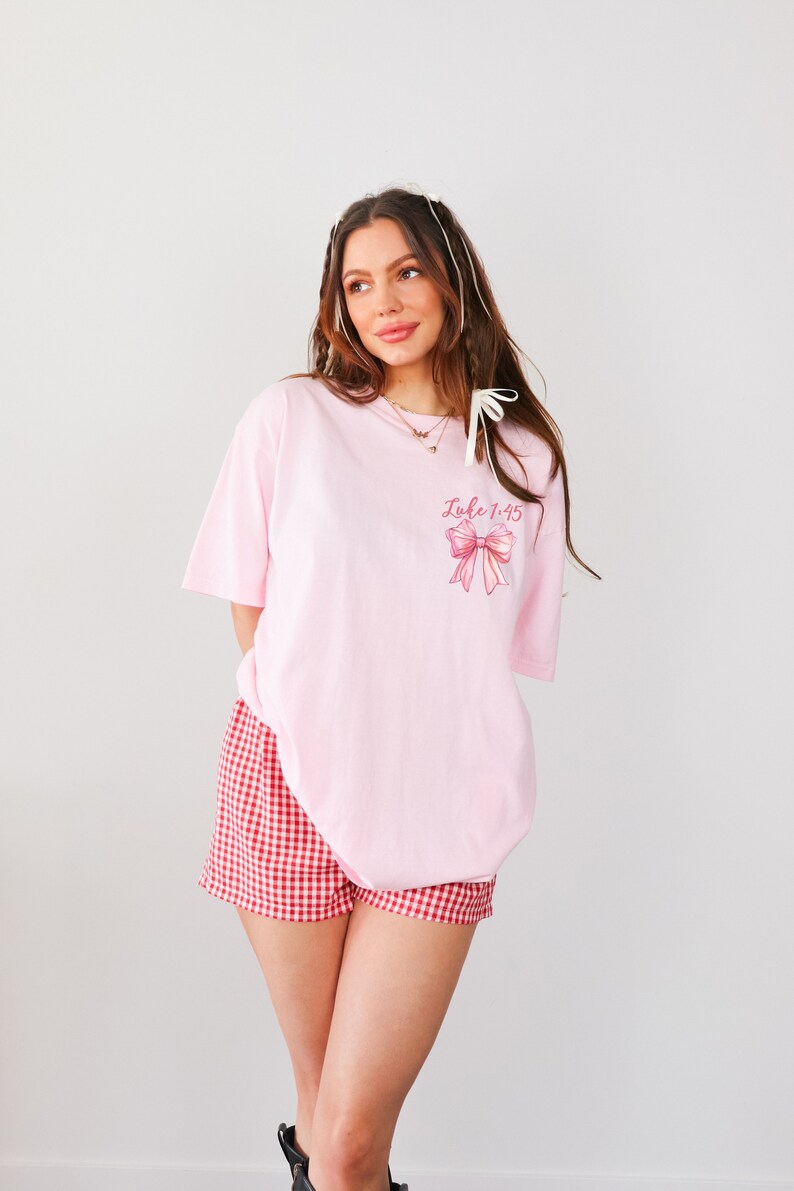 May include: An oversized pink t-shirt with a pink bow and the text "Luke 1:45" printed on the front. The model is wearing red and white gingham shorts.