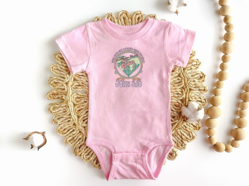 K&ouml;nnte beinhalten: Rosaer Baby-Strampler mit einem Grafikdruck eines herzf&ouml;rmigen Globus mit den Worten "Gott hat die Welt so sehr geliebt" und "Johannes 3:16" darauf.