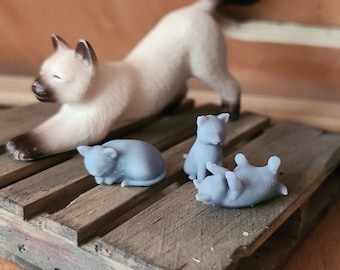 May include: A white Siamese cat figurine stretches out on a wooden platform. Three smaller grey cat figurines are lying on the platform, one is curled up sleeping, one is sitting up, and one is lying on its back.