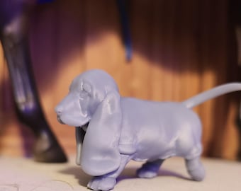 May include: A light blue basset hound figurine with long ears and a short tail. The dog is standing on a light-coloured surface, with a blurred background of a wooden structure and a dark object.