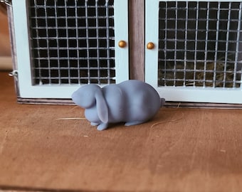 May include: A small, grey, ceramic rabbit figurine. The rabbit is lying on its side with its head turned to the left. The rabbit has a long, floppy ear and a short, stubby tail.