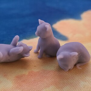 Op de afbeelding: Drie kleine, paarse, 3D-geprinte kattenfiguren. E&eacute;n kat ligt op zijn rug, een andere zit rechtop en de derde is opgerold.