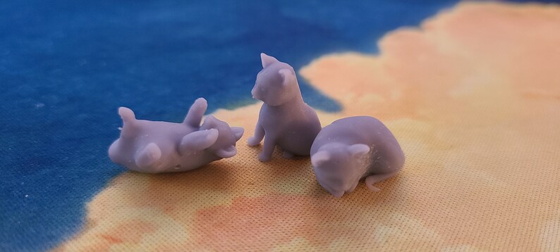 Op de afbeelding: Drie kleine, paarse, 3D-geprinte kattenfiguren. E&eacute;n kat ligt op zijn rug, een andere zit rechtop en de derde is opgerold.