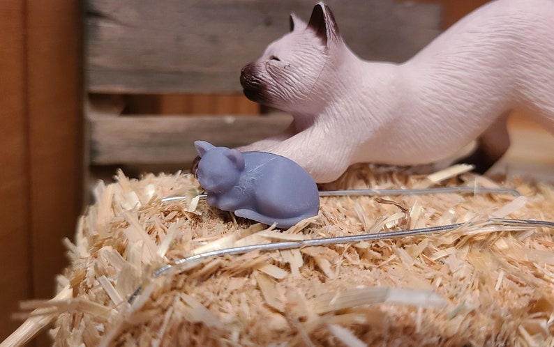 Op de afbeelding: Een klein, grijs, slapende kattenbeeldje op een bed van stro met een groter wit kattenbeeldje op de achtergrond.