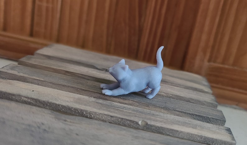 Op de afbeelding: Een klein, grijs, 3D-geprint beeldje van een kat in een speelse pose. De kat ligt op zijn buik met zijn voorpoten gestrekt en zijn staart opgerold.