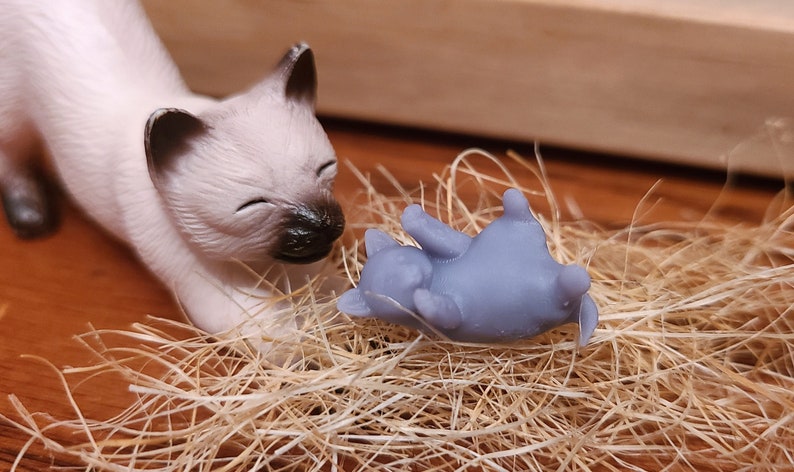 Op de afbeelding: Een wit Siamese kattenbeeldje met gesloten ogen, kijkt neer op een kleiner grijs kattenbeeldje dat op zijn rug ligt. Beide beeldjes liggen op een bed van bruin stro.