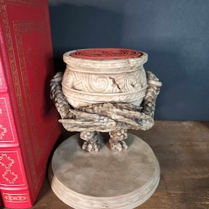May include: A ceramic bookend in the shape of a creature holding a pot. The pot is decorated with a swirling pattern and has a red top. The creature has a detailed, textured surface and is standing on a round base.