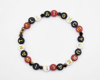 May include: A beaded bracelet with the words "ALL AT ONCE" spelled out in gold letters on black, red, and white round beads. The bracelet is on a white background.