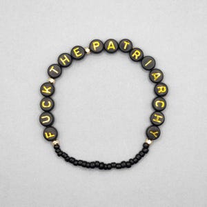 May include: Black beaded bracelet with the words "FUCK THE PATRIARCHY" spelled out in gold letters on black circular beads. The bracelet is on a light gray background.
