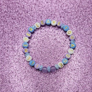 May include: A bracelet made of colourful beads, spelling out "NEVER LEAVE YOU ALONE" in gold letters. The beads are in shades of blue, purple, and white, with clear and purple accent beads.