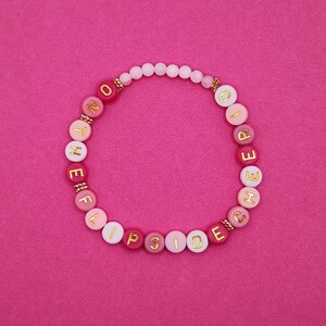 May include: A pink beaded bracelet with the words "NOT THE OTHER SIDE" spelled out in gold letters. The bracelet features a mix of pink and white beads against a pink background.