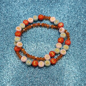 May include: Two beaded bracelets with the words "EVERYONE'S BOUND BOTH HERE" and "THEY'RE NOT" spelled out in gold letters on colorful beads. The bracelets are on a sparkly blue surface.