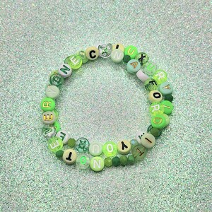 Puede incluir: Una pulsera circular de cuentas en varios tonos de verde, incluyendo lima, esmeralda y menta. La pulsera está compuesta por cuentas redondas de plástico, algunas translúcidas y otras opacas, con letras negras y doradas que forman la frase.