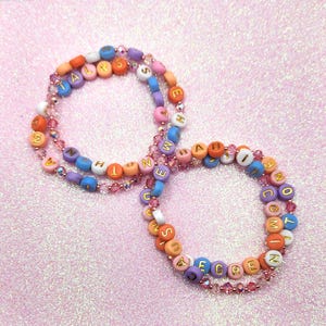 May include: Two beaded bracelets with colorful letter beads spelling out words. The bracelets are made of round beads in various colors, including orange, blue, purple, and pink. The letters are gold. The bracelets are on a pink glittery surface.