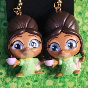 May include: A pair of dangle earrings featuring cartoon-like figures with brown hair, blue eyes, and green dresses. Each figure holds a pink teacup. The earrings have gold-colored hooks.