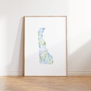 May include: A framed art print featuring the outline of Delaware, filled with abstract blue and green line art. The artwork is displayed on a white wall, with a light wooden frame and a wooden floor.