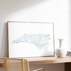 Op de afbeelding: Een ingelijste kunstdruk met een gestileerde kaart van North Carolina, weergegeven in blauwe en groene tinten. Het kunstwerk wordt gepresenteerd op een houten bureau, samen met een witte vaas en een stoel met een geweven rugleuning. De print zit in een licht houten frame.