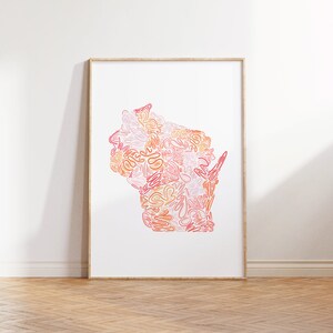 May include: A framed art print featuring the outline of Wisconsin filled with abstract, swirling lines in shades of pink, orange, and peach. The artwork is displayed on a white wall above a wooden floor.