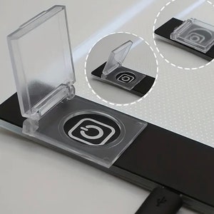 Op de afbeelding: Een zwarte tekentablet met een doorzichtige plastic afdekking over de aan/uit-knop. De afdekking is scharnierend en kan worden opengeklapt om de aan/uit-knop te onthullen.