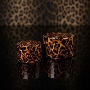 May include: Two cylindrical herb grinders featuring a brown and black leopard print pattern. The grinders are stacked in multiple sections, showing a glossy finish and a consistent animal print design across both the smaller and larger units.