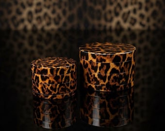 May include: Two cylindrical herb grinders featuring a brown and black leopard print pattern. The grinders are stacked in multiple sections, showing a glossy finish and a consistent animal print design across both the smaller and larger units.