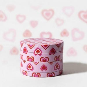 May include: A cylindrical metal herb grinder featuring a repeating pattern of pink, red, and dark red hearts. The grinder consists of three stacked circular sections, each decorated with the same heart motif, set against a light background.