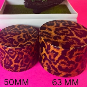 May include: Two cylindrical containers with lids, featuring a leopard print pattern in shades of brown and orange. The smaller container is labeled "50MM" and the larger one is labeled "63 MM". The background is a bright pink.
