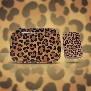 May include: A rectangular metal rolling tray and a smaller matching rectangular metal tin, both featuring a repeating brown and black leopard print pattern against a tan background with rounded corners and raised edges.