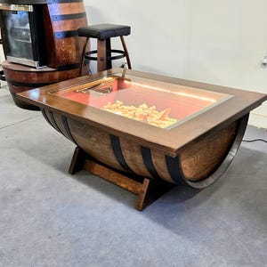 May include: A handcrafted coffee table made from a repurposed wooden barrel. The table has a rectangular glass top with a wooden frame, revealing a red interior and decorative elements. The barrel is supported by wooden legs and has black metal bands.