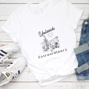 Puede incluir: Camiseta blanca con un diseño gráfico negro de una escena del desierto con cactus y el texto "Unleash the Extraordinary".