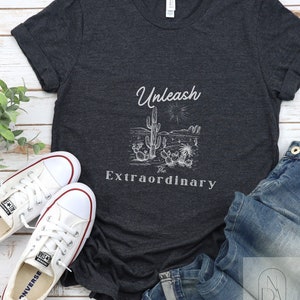 Puede incluir: Una camiseta gris oscuro jaspeada con un gráfico blanco de una escena del desierto con cactus y el texto "Unleash the Extraordinary".