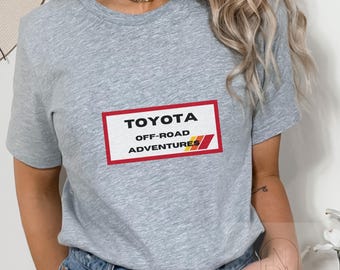 Op de afbeelding: Grijs T-shirt met korte mouwen met een rode rechthoekige afbeelding met de tekst "TOYOTA OFF-ROAD ADVENTURES" in witte letters.