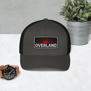 Puede incluir: Gorra de camionero gris oscuro con la parte trasera de malla negra. La parte delantera presenta un parche rectangular con un diseño de sol rojo y la palabra "OVERLAND" en blanco. Un reloj de pulsera con una correa de cuero marrón está en primer plano.