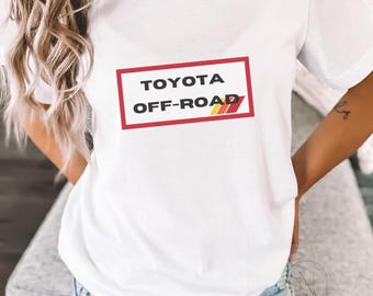 Op de afbeelding: Wit T-shirt met een rode box en de tekst "TOYOTA OFF-ROAD" in zwarte letters met een kleurrijk streepontwerp.