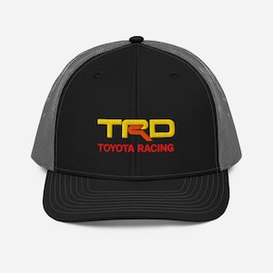 Op de afbeelding: Een zwarte en grijze truckercap met een rood, geel en oranje geborduurd logo met de tekst "TRD TOYOTA RACING".