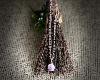 Op de afbeelding: Een zilveren ketting met een lichtpaarse kristallen hanger. De ketting hangt aan een bruine bezem.