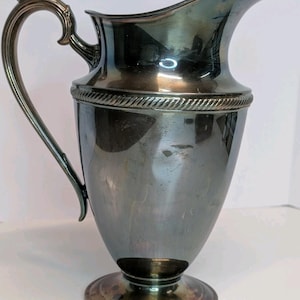 May include: A silver-colored pitcher with a curved handle and a spout. The pitcher has a decorative rope-like design around the top and sits on a round base. The handle has an ornate design at the top.
