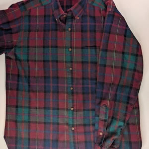 May include: A long-sleeve button-down shirt featuring a plaid pattern in shades of burgundy, dark green, and navy blue. The shirt has a button-down collar, a single chest pocket, and brown buttons. The inner collar label displays the text LOBO.