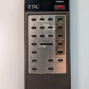 May include: Vintage Zenith Z-TAC remote control. The black remote has silver buttons for channel, volume, and other functions. The text "ZENITH ADDRESSABLE CONTROL SPACE COMMAND" is printed on the top.