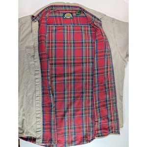 May include: A tan long-sleeve button-down shirt with a red, blue, and green plaid flannel lining. The shirt features a Cabela's brand label at the inner collar, a front button placket, and a chest pocket on the left side of the tan exterior.