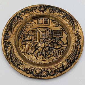 May include: A circular brass decorative plate featuring a raised relief scene of a group of people gathered outside a building. The border is decorated with intricate scrollwork and leaf patterns, with a dark patina highlighting the details.