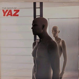 Peut inclure: Un boîtier CD présentant la pochette de l'album The Best of Yaz. La pochette montre deux mannequins lisses et sans traits, de couleur claire, devant un mur à panneaux horizontaux blancs. Le texte THE BEST OF YAZ est imprimé en rouge et gris.