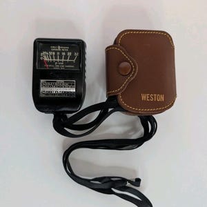 May include: A black Weston exposure meter with a white dial and numerical scales, featuring a black neck strap and a brown leather protective case with the word WESTON embossed on the front. The device includes a chart for still and cine cameras.