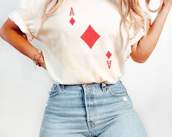 May include: Ivory-colored t-shirt featuring a red Ace of Diamonds playing card design. The shirt has short sleeves and a relaxed fit. The word "IVORY" is printed in the upper left corner.