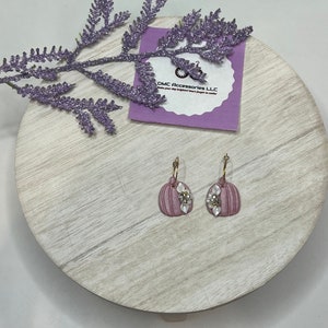 May include: A pair of pink pumpkin earrings with white pearl accents. The earrings are on a wooden surface with a purple flower sprig.