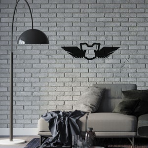 May include: A black metal wall decoration in the shape of a shield with wings, mounted on a grey brick wall. The artwork is above a grey sofa with cushions and a dark blanket. A black floor lamp and a coffee table with books and glassware are in the foreground.