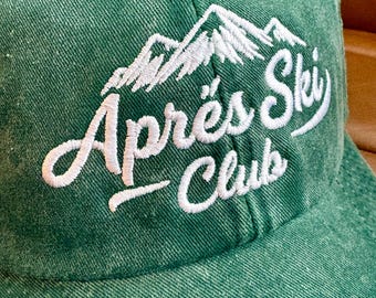 Puede incluir: Gorra de béisbol verde con las palabras "Après Ski Club" bordadas en blanco sobre un diseño de cordillera. La gorra tiene una visera curva y un botón en la parte superior.
