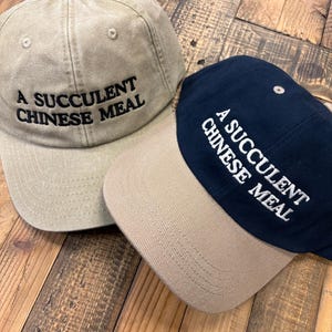 Puede incluir: Dos gorras de b&eacute;isbol con el texto "A succulent Chinese meal" bordado en la parte delantera. Una gorra es beige y la otra es azul marino.